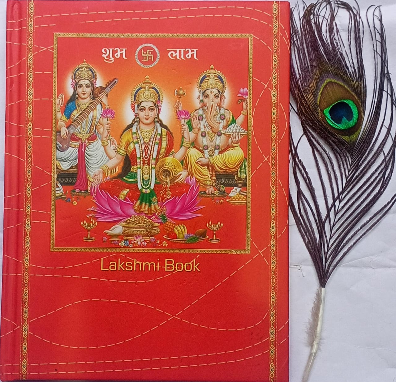 Red book titled 'Lakshmi Book' with an illustration of deities on the cover, accompanied by a peacock feather.
